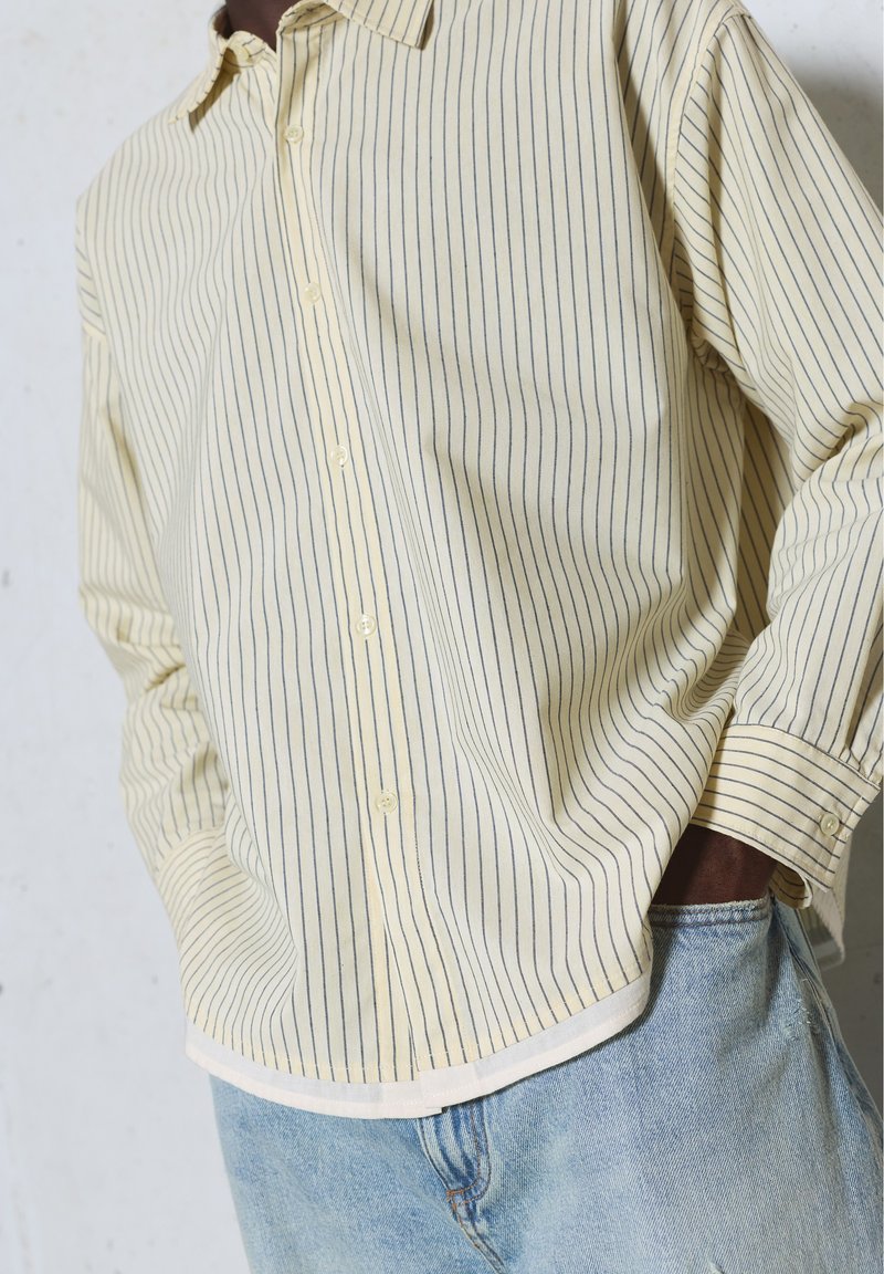Personne portant une chemise beige à manches longues avec de fines rayures verticales et un jean en denim bleu clair, une main dans la poche.