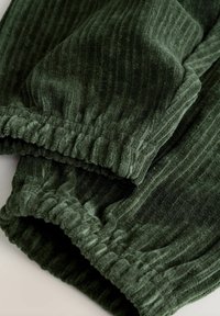 Tummanvihreät corduroy-housut, joissa on elastiset kootut hihnat, näyttäen tekstuurisia pystyraitoja ja pehmeitä kankaankappaleita.
