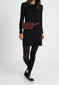 Svart långärmad klänning, knälång, i kombination med svarta leggings och sneakers. Röd bälteväska sitter vid midjan, enkel design.