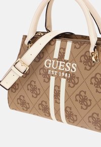 Sac à main à motifs marron avec des accents crème, quincaillerie dorée, double anses et une bandoulière amovible. Porte le logo "GUESS EST 1981".