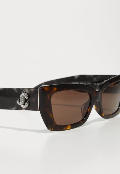 Lunettes de soleil écaille de tortue avec un design carré épais et des verres marron. Dotées d'un détail décoratif en argent sur une branche noire.