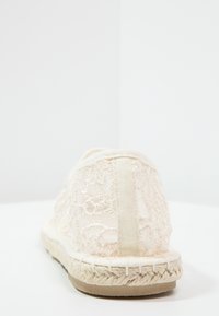 Espadrille à dos en dentelle crème, présentant des motifs floraux complexes, un bord en corde et une tige en textile doux pour le confort.