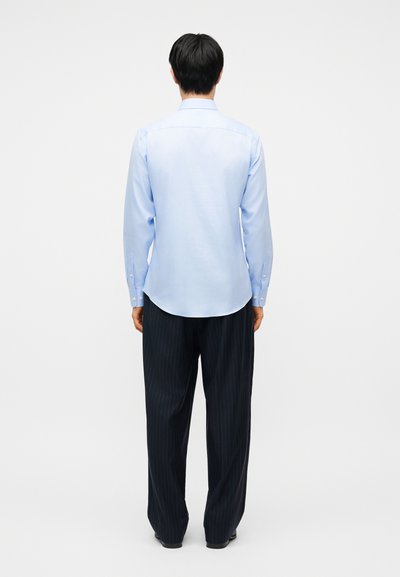 Άνδρας που στέκεται γυρισμένος προς τα πίσω, φοράει ανοιχτό μπλε πουκάμισο με μακριά μανίκια και φαρδιά σκουρόχρωμα παντελόνια με στενές ρίγες σε μονόχρωμο φόντο.