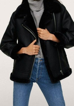 Personne portant une veste en peau lainée noire avec col et poignets duveteux, pull à col roulé gris et jean bleu, tenant la veste ouverte à la fermeture éclair.