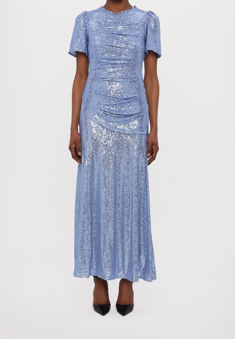 Femme portant une robe longue bleu clair, à manches courtes, ornée de sequins, associée à des talons noirs à bouts pointus.