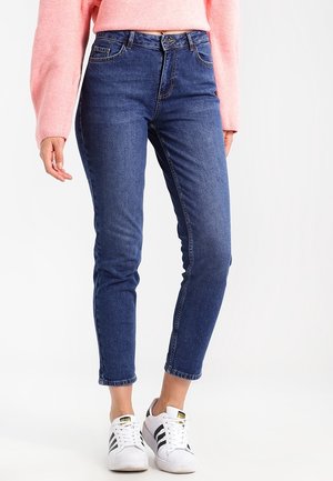 Personne portant un jean slim bleu, un pull rose et des baskets blanches avec des rayures noires, debout sur un fond blanc uni.