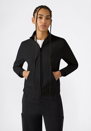 Jong persoon met gevlochten haar, gekleed in een zwart jack met ritssluiting en zwarte broek, staand met handen in de zakken van het jack tegen een witte achtergrond.