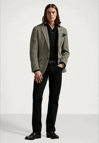 Ung man med axellångt brunt hår, iklädd en grå kavaj, svart polotröja, svarta byxor och svarta dressade skor, står med ena handen i fickan.