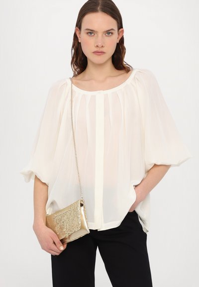 Chemisier blanc, transparent, avec des manches bouffantes et des plis sur le devant. Associé à une petite pochette en métal doré avec une bandoulière en chaîne. Pantalon noir.