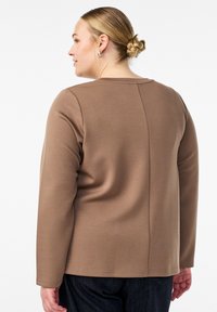 Brązowa bluzka z długim rękawem o gładkiej teksturze, okrągłym dekolcie i widocznych detalach szwów z tyłu. Uzupełniona ciemnymi dżinsami.