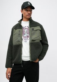 Chaqueta de forro polar en verde oscuro con una superficie texturizada, que cuenta con un cierre frontal, un bolsillo en el pecho y detalles de paneles de tela contrastantes.