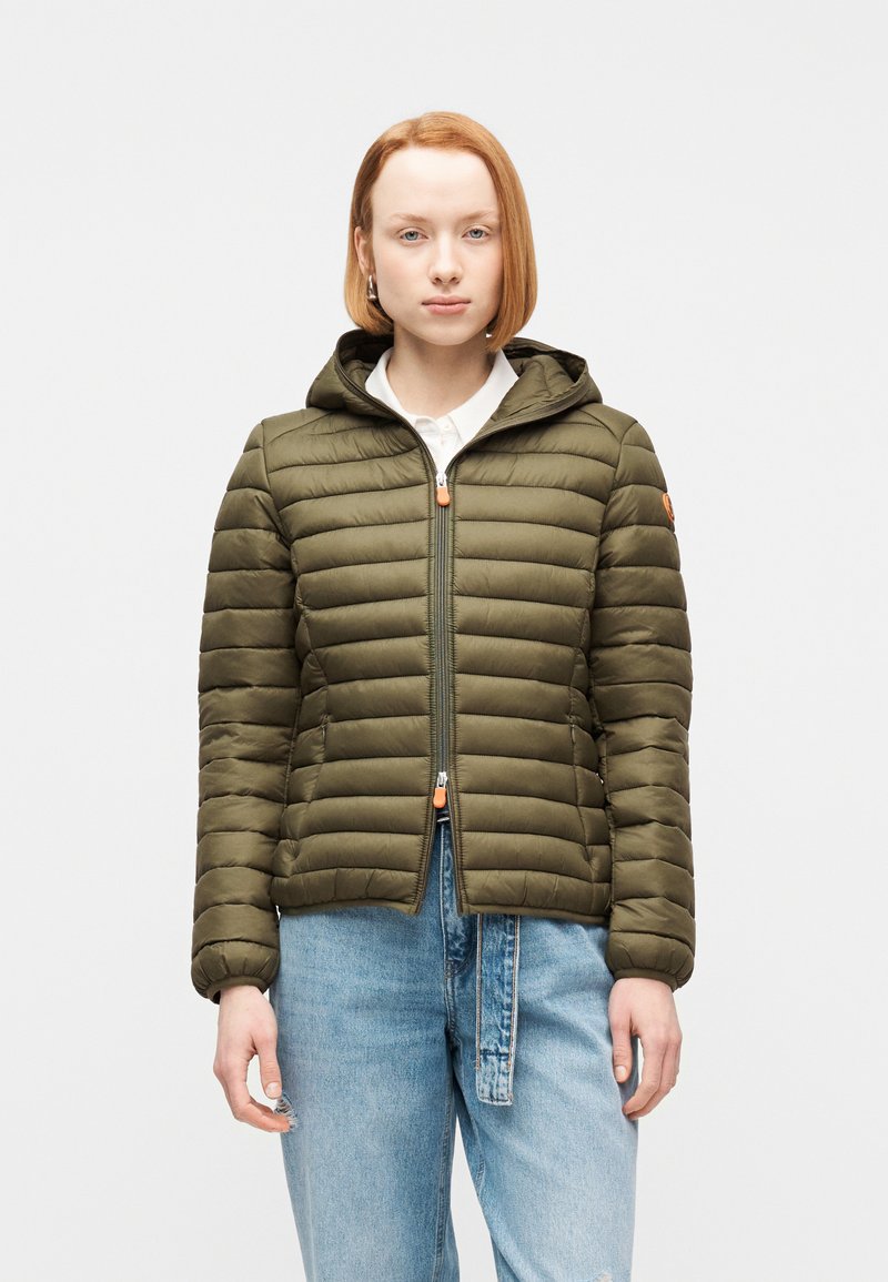 Νεαρό άτομο με κοντά κόκκινα μαλλιά που φοράει μια χακί καπιτονέ μπουφάν με κουκούλα και ανοιχτόχρωμο τζιν, μπροστά από απλό φόντο.