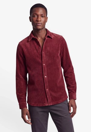 Man wearing a long-sleeve, button-up red corduroy shirt and dark pants, standing against a plain white background.