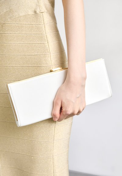 Pochette rectangulaire blanche avec une surface lisse et un fermoir doré, tenue dans une main contre une robe texturée de couleur claire.
