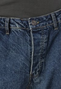 Σκούρα μπλε τζιν παντελόνι με κούμπωμα κουμπιού, διαθέτοντας υφή υφάσματος και ορατές ραφές κατά μήκος των ραφών.