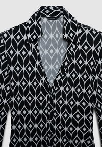 Schwarz-weiße Bluse mit geometrischem Rautenmuster, ausgestattet mit einem Kragen, durchgehender Knopfleiste und glatter Stofftextur.