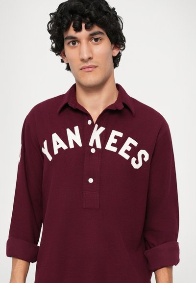 Burgunderfarget langermet polo skjorte laget av mykt stoff. Har hvit "Yankees" tekst, knappestolpe og oppbrettede ermer.