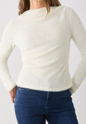 Kobieta ubrana w dopasowany, długi biały sweter z miękkiego materiału, zestawiony z ciemnoniebieskimi, wysokim stanem jeansami, ręce oparte na biodrach.