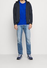 Granatowa bluza z zamkiem i kapturem na sznurku, niebieska koszulka z okrągłym dekoltem, jasnoniebieskie dżinsy oraz granatowe buty sportowe z białymi akcentami.