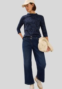 Top a maniche lunghe blu navy con stampa zebra, jeans a gamba larga in denim scuro e borsa beige. Completato da sneakers beige e un cappellino chiaro.