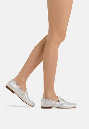 Woman’s legs wearing silver leather loafers with low wooden heels, walking against a plain light gray background.