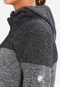 Hættetrøje i tekstureret sort og gråt materiale med et mønstret design og en lille logo på nederste højre side.