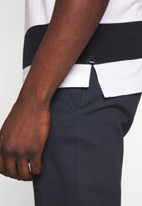 Polo rayé blanc et marine, avec un détail d'ourlet fendu. Un pantalon foncé au toucher lisse complète la tenue. Main positionnée sur le côté.