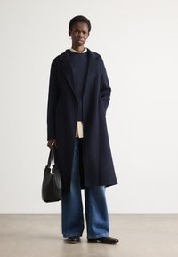 Mulher vestindo um casaco longo azul-marinho, suéter escuro, jeans azul de perna larga, sapatos pretos planos, carregando uma bolsa preta, de pé contra um fundo claro e simples.
