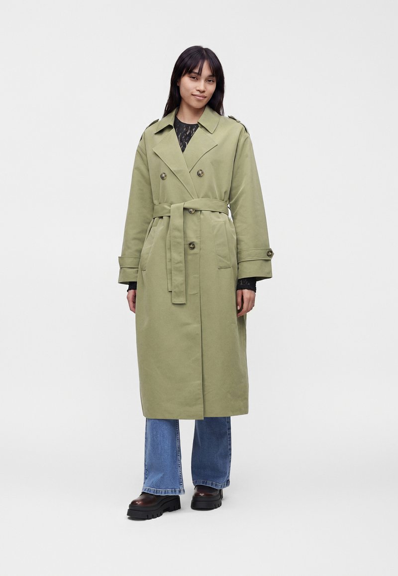 Femme portant un long trench-coat ceinturé vert clair sur un jean bleu et des chaussures noires épaisses, debout devant un fond uni clair.