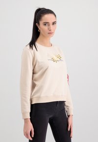 Béžová mikina s okrúhlym výstrihom, ktorá má zlaté logo a čierny text. Vyrobená z hladkého materiálu, má rebrované manžety a voľný strih.
