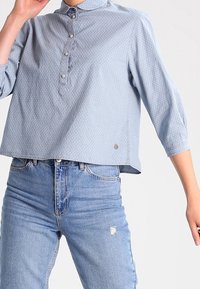 Ljust blå knapp-up skjorta med krage, trekvartsärmar, texturerad tyg och vita prickar, kombinerad med slitna denimjeans.