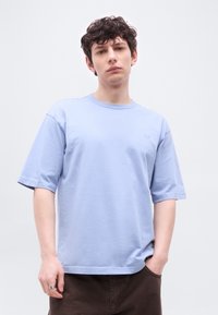 Νέος άνδρας με σγουρά μαλλιά που φοράει μια ανοιχτό μπλε πουκαμίσα σε μεγάλο μέγεθος και σκούρο παντελόνι, στέκεται με το ένα χέρι στην τσέπη μπροστά σε λευκό φόντο.