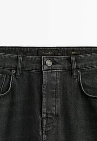 Mustat denim-housut kapealla istuvuudella, joissa on metallipainike, viisi taskua ja hienovarainen tekstiuri. Tägki näkyy vyötäröllä.