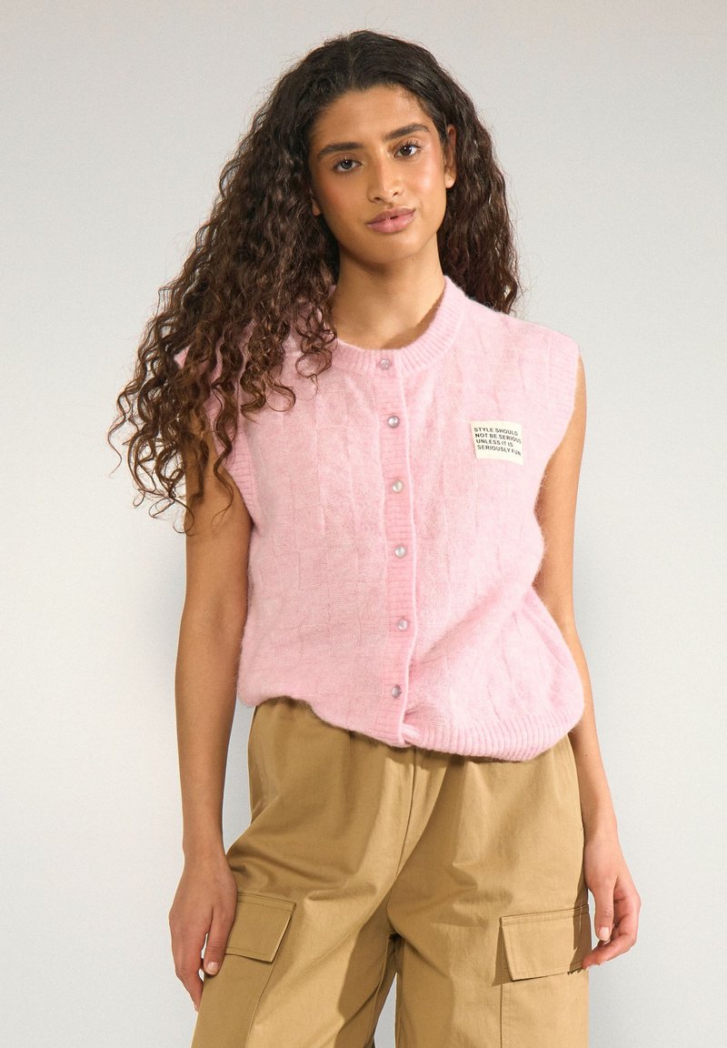 Femme aux longs cheveux bouclés portant un gilet rose sans manches boutonné et un pantalon cargo beige, debout devant un fond uni.