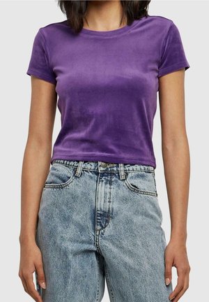 Vrouw draagt een aansluitende paarse fluwelen crop top met korte mouwen, gecombineerd met een lichtblauwe high-waisted acid-wash jeans.