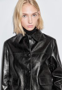 Zwarte leren jas met een glanzende afwerking, voorzien van een knoopsluiting aan de voorkant, een kraag, en twee voorzakken. Getextureerd en gestroomlijnd ontwerp.