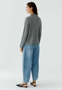 Pull en maille gris avec un col haut, associé à un jean en denim bleu clair à coupe ample et des chaussures noires à bride arrière. Il y a un petit détail au dos.