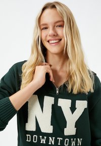 Sudadera de color verde oscuro con cuello con cremallera. Presenta un gran texto en color crema "NY" y la palabra "DOWNTOWN" debajo. Puños y dobladillo de canalé.