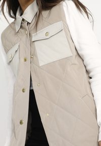 Hellbeige gesteppte Weste mit spitzem Kragen, zwei Brusttaschen und goldenen Hardware-Details. Strukturiertem Stoff mit subtiler Glanz.