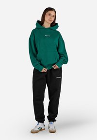 Sweatshirt verde com capuz e bolso frontal, combinado com calças de moletom pretas com um logótipo. Usado com ténis brancos e cinzentos com riscas.