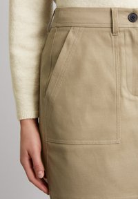Jupe kaki en tissu durable avec une finition texturée, dotée d'une poche avant et d'une fermeture à bouton noir. Design de taille lisse.
