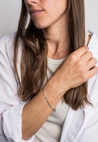 Sidabrinė grandinėlė apyrankė, dėvima ant riešo, su sujungtais segmentais ir maža žaviu. Rankos poilsio padėtis, vidutinio odos tono.