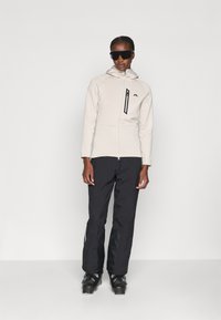 Lichtgekleurde, met rits afsluitbare fleece jas met een capuchon en zwarte accenten, gecombineerd met zwarte skibroeken en zwarte skischaatsen. Accessoires omvatten een zonnebril.
