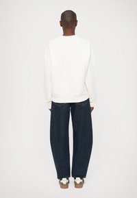 Felpa bianca a maniche lunghe, abbinata a jeans in denim scuri a gamba larga. Le scarpe da ginnastica presentano dettagli verdi e il logo sul tallone.