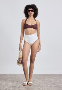 Zweiteiliges Badebekleidung bestehend aus einem bordeauxfarbenen strukturierten Bralette und einem hochgeschnittenen weißen Bikiniunterteil. Accessoires umfassen eine geflochtene Tasche und weiße Flip-Flops.
