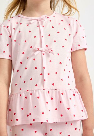 Top rosa chiaro a maniche corte con motivo a cuori rossi, con due piccoli fiocchi frontali e orlo a peplo, indossato da un bambino.