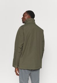 Olive green jacket with a high collar, made of smooth fabric. Features straight cut and subtle seam details, paired with gray pants.