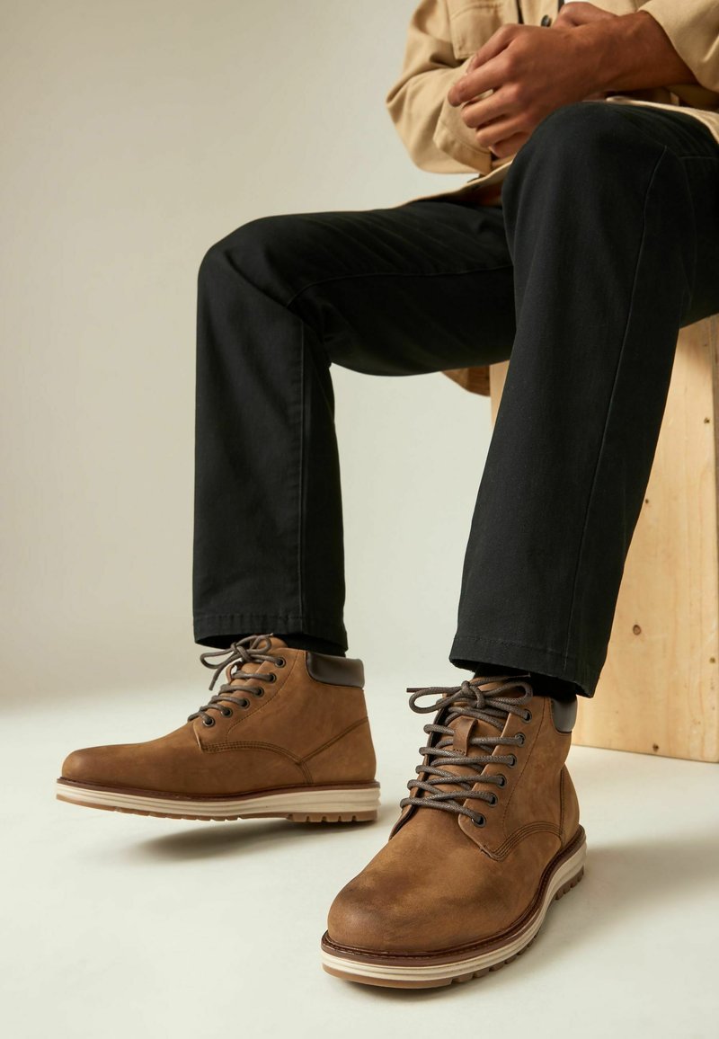 Bruine leren enkelboots met bruin vetersluiting, beige tussenzool en textuur. Gecombineerd met zwarte broek voor een lichte achtergrond.