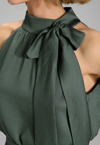 Donkergroen mouwloos blouse met een hoge kraag, voorzien van een grote strik bij de hals en een gestructureerde stof.