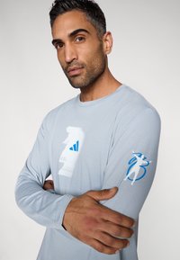 Light gray long-sleeve shirt with a white and blue graphic logo on the front and blue accent on the sleeve, made from soft material.